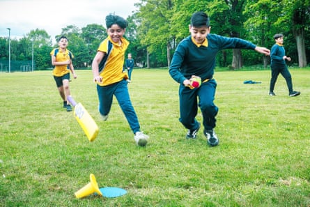 Two young Asian boys race for a cricket wicket; one has thrown the bat towards it while the other runs with the ball. Other boys are in the background.