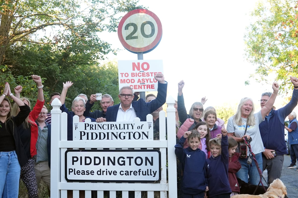 People of all ages including children plus a dog stand by a sign reading Principality of Piddington above the village sign and a white gate.