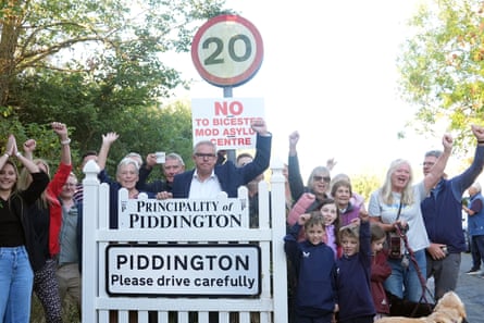 People of all ages including children plus a dog stand by a sign reading Principality of Piddington above the village sign and a white gate.