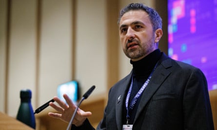 Mustafa Suleyman fala aos repórteres na cúpula atrás de um microfone e na frente de uma tela; ele usa uma jaqueta escura e um suéter preto com gola polo