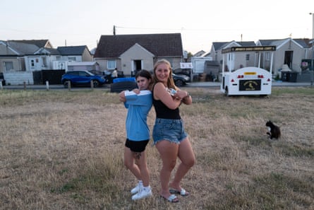 Two girls pose back to back, arms crossed, on a grassy patch in front of houses