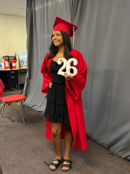 A teenager in a red cap and gown, black dress and black sandals smiles while posing with white letters that says “26”.