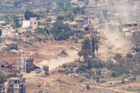 A vehicle drives past a partially destroyed building with an Israeli flag displayed on the side, with a general view of the surrounding village showing extensive damage from bombings.