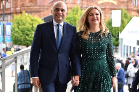 Javid in a navy blue suit and tie and and his wife, Laura wearing a green spotted dress walking toward the camera.
