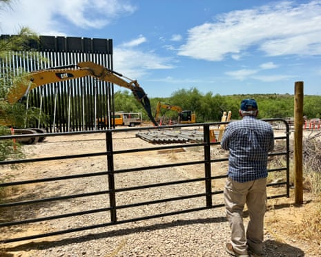 a man stands behind a gate and looks at construction equipment and a partially built border fence