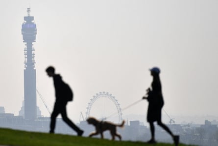 Two people walk a dog in a hilltop with a view of London behind showing the BT tower and London Eye