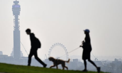Two people walk their dog with the BT Tower and the London Eye seen in the hazy background at Primrose Hill in north London