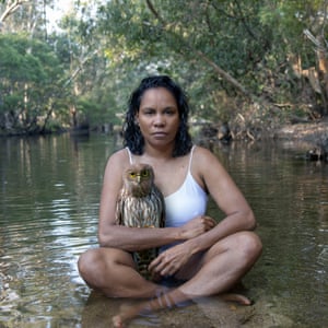 A woman sits in the shallows of a river bank holding a pet owl.