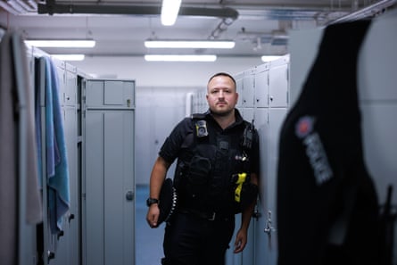 Daniel Bragg stands in a locker room at Ashton police station, leaning against grey metal lockers. He is quite young with very short dark hair and beard. He holds his cap under his arm.
