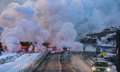 Smoke billowing from orange and black molten lava which is flowing over a road with emergency vehicles parked on it and snow-covered fields beside it