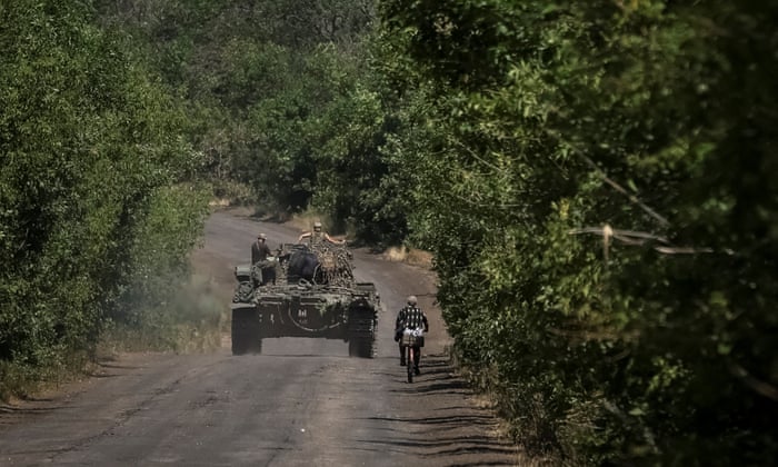 Ukrainian servicemen ride on a military vehicle as a local resident rides a bicycle in the Donetsk region