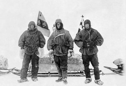 A balc and white photograph of three men in old fashoned cold weather gear stand in front of loaded sleds and flags.