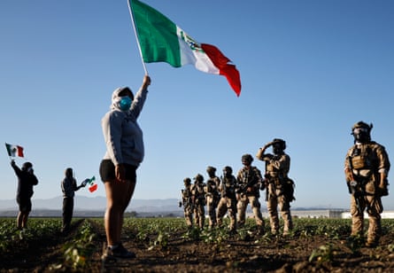 three protesters wearing masks hold California flags and stand facing a line of federal agents in military clothing