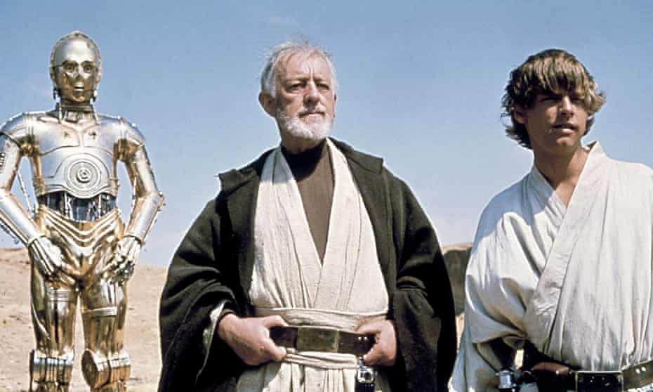 C3-P0 (Anthony Daniels), Obi-Wan Kenobi (Alec Guinness) and Luke Skywalker (Mark Hamill) stand against a blue sky, looking into the distance in a desert.