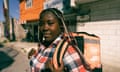 Berline Augustin, a black woman with long braids, wears a chequered shirt and carries an orange food-delivery rucsac on her back. Behind her the walls are made from concrete blocks.
