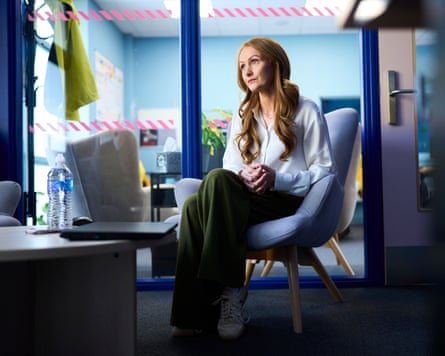 Lauren Monks sits on a blue chair with her legs folded, in front of a glass wall with bright blue frames. She has long wavy reddish-blond hair and wears a white shirt and green trousers.