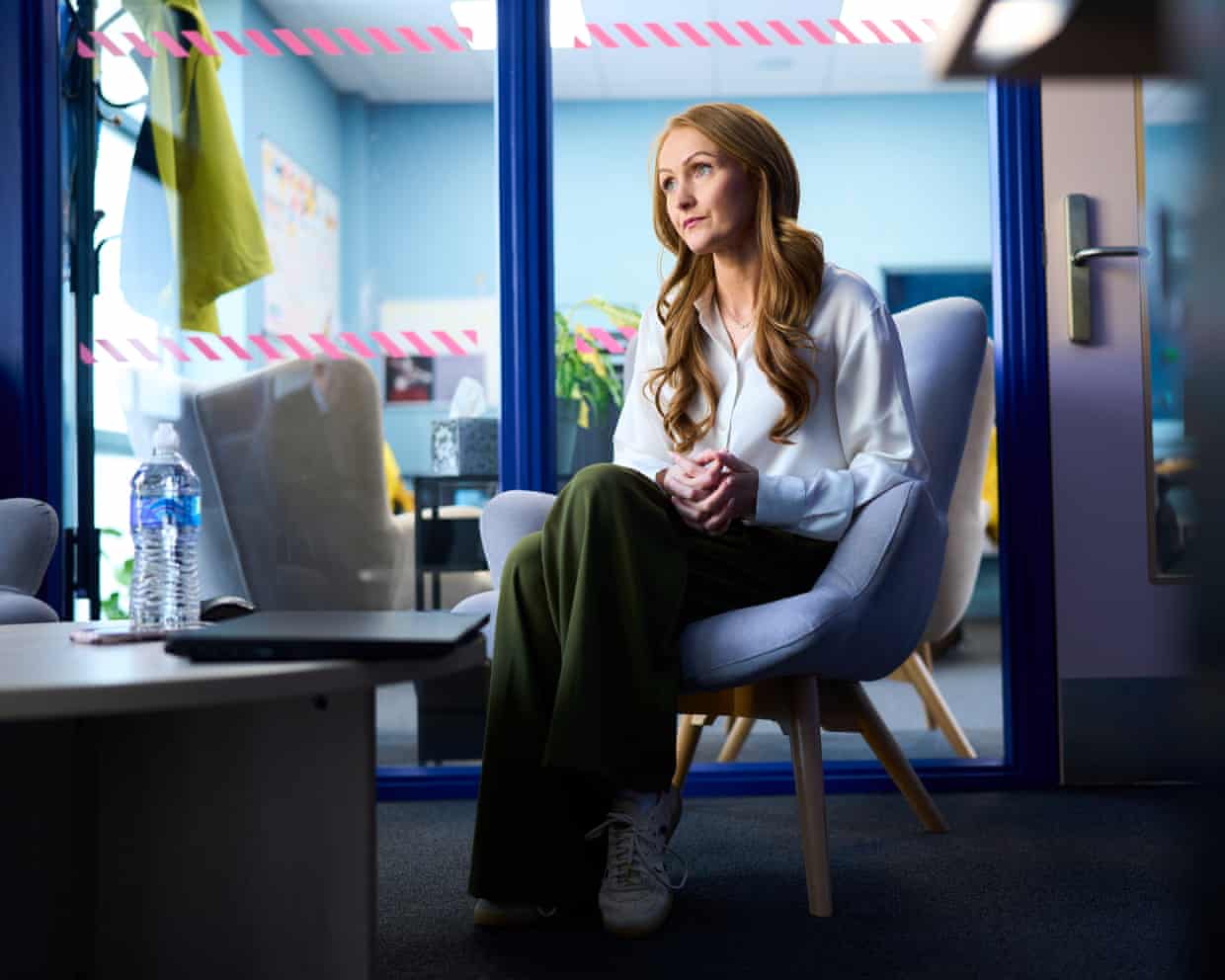 Lauren Monks sits on a blue chair with her legs folded, in front of a glass wall with bright blue frames. She has long wavy reddish-blond hair and wears a white shirt and green trousers.