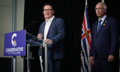 One man wearing a blue suit speaks at a podium marked with a sign saying 'Conservative party of British Columbia,' while another man wearing a navy suit stands by