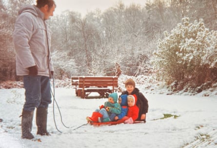 A man stands in a snowy field holding a rope for a sledge carrying toddler triplets, with a young boy crouched down behind them