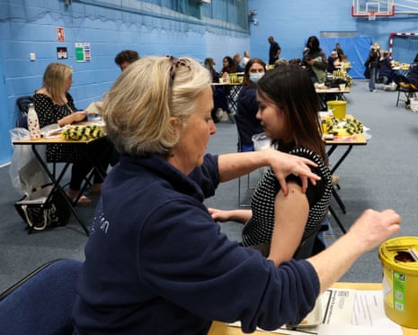 A healthcare worker administers a vaccination to a young woman at a table in a sports hall