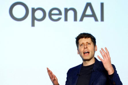 A man gestures while speaking in front of a screen that says ‘OpenAI’