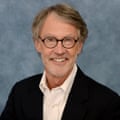 Smiling man with round glasses and blazer sits for portrait photo.