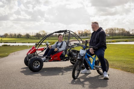 A woman in a red off-road buggy and a man on a small blue off-road bike with large lawns in the background