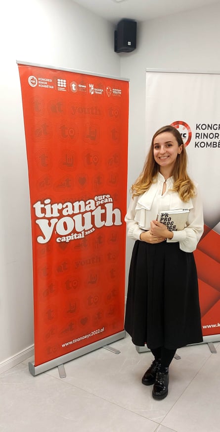 Dafina Peci smiling for a photograph standing next to a large orange banner saying Tirana Youth.