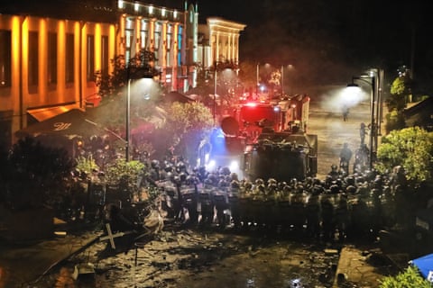 A line of riot police with shields and vehicles in front of a grand stone building with pillars.