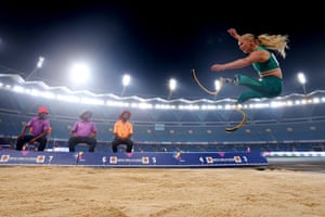 Three judges, two in purple polo shirts and one in a orange polo shirt, and hats sit next to long jump pit as an athlete with race blades and wearing the green of Australia leaps through the air inside a flood-lit stadium