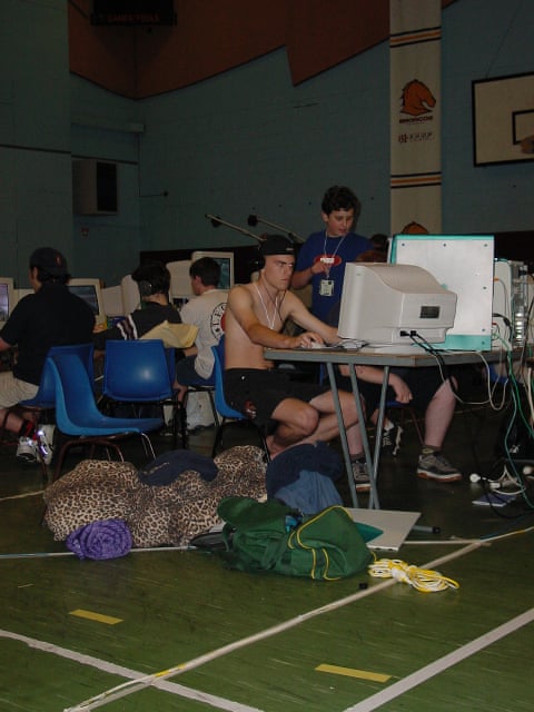 A group of young men sitting at tables in a school hall with assorted computers on the desks and clothes and sleeping bags on the floor