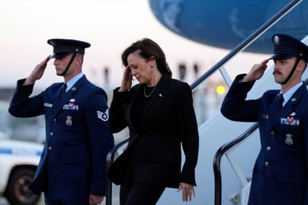 Harris, wearing a black suit, salutes as she steps down the stairs of a plane, as two Air Force men in uniform stand between her