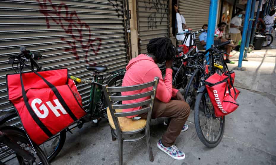 People sit in chairs next to businesses with their storefront grates closed, surrounded by bicycles bearing Grubhub branded bags.