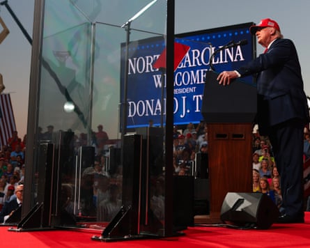 Aa man in suit wearing red baseball cap, speaks at lectern in front of crowd as large screen beside him displays âÂÂNorth Carolina welcomes President Donald J TrumpâÂÂ