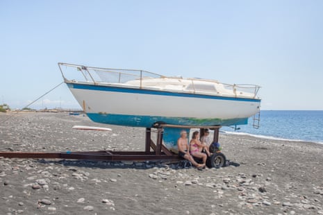 A German family sits in the shade of a boat pulled aground on the beach in Stromboli, a volcanic island of Sicily’s Aeolian archipelago. Italy, 2022