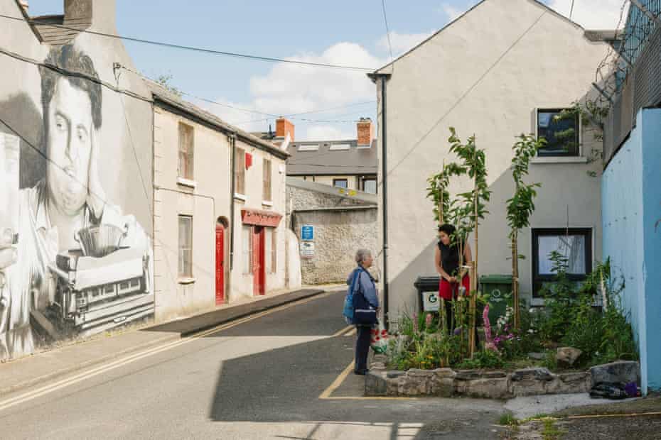 Wide shot of the parklet and the surrounding street. Two women talking. Wall on left has giant mural showing writer Brendan Behan with typewriter.
