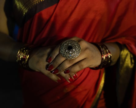 A woman’s hands, wearing large rings and gold bangles, clasped in front of her red sari.