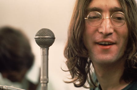 John Lennon, with long hair and round wire-framed glasses, smiles as he sings next to a microphone in the film Let It Be