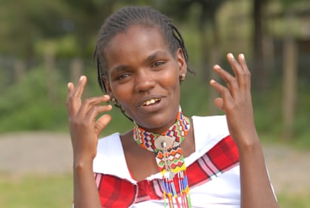 A young African woman with a beaded necklace smiles and gestures at the camera