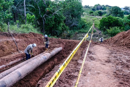 Two men with spades dig in a ditch that carries a wide pipeline as others, also in hard hats, work in the distance