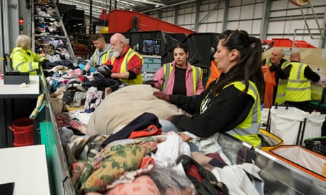 A line of people in a factory inspect and gather large piles of clothing coming down a conveyor belt