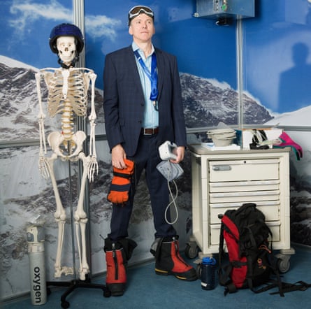 Hugh Montgomery in a suit, ski goggles and boots, with medical equipment in his hand, a skeleton and oxygen mask one side of him and a rucksack on the other