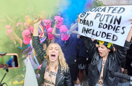 Protesters, some wearing only a leather jacket with slogans painted on their chests, and others wearing pink balaclavas, raise their fists, shout and hold placards
