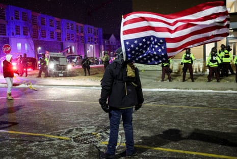 A demonstrator, dressed in dark winter gear, stands before a gaggle of law enforcement officers in high-vis jackets while an upside down American flag, strapped to his back, waves in the wind.