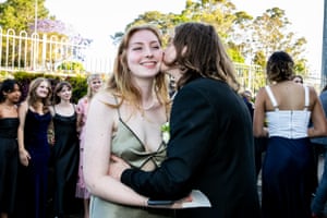 A young person kisses another on the cheek while behind them girls pose for photos in their formal gowns