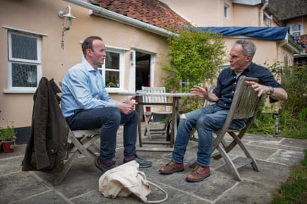 Richard Route, in blue shirt and jeans, sits talking to Julian Calderara, who is raising a hand as he makes a point. They are sitting on wooden chairs in the back garden.