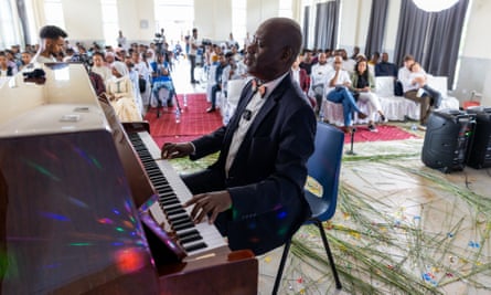 Girma Yifrashewa, wearing a navy suit and colorful bow tie, sits on a plastic chair and plays at a wooden piano in the foreground before an audience at the Homosha school for girls
