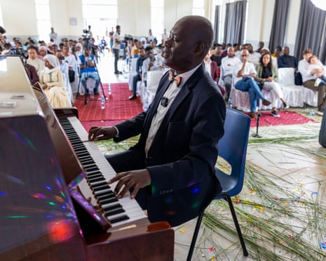 An African man in a bow tie and suit sits at a grand piano with his eyes closed as an audience listens