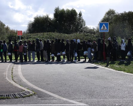 Migrants queue for food handouts, holding plastic bags, near trees in Calais