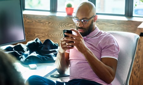 Man with beard and glasses sitting at desk, checking smartphone